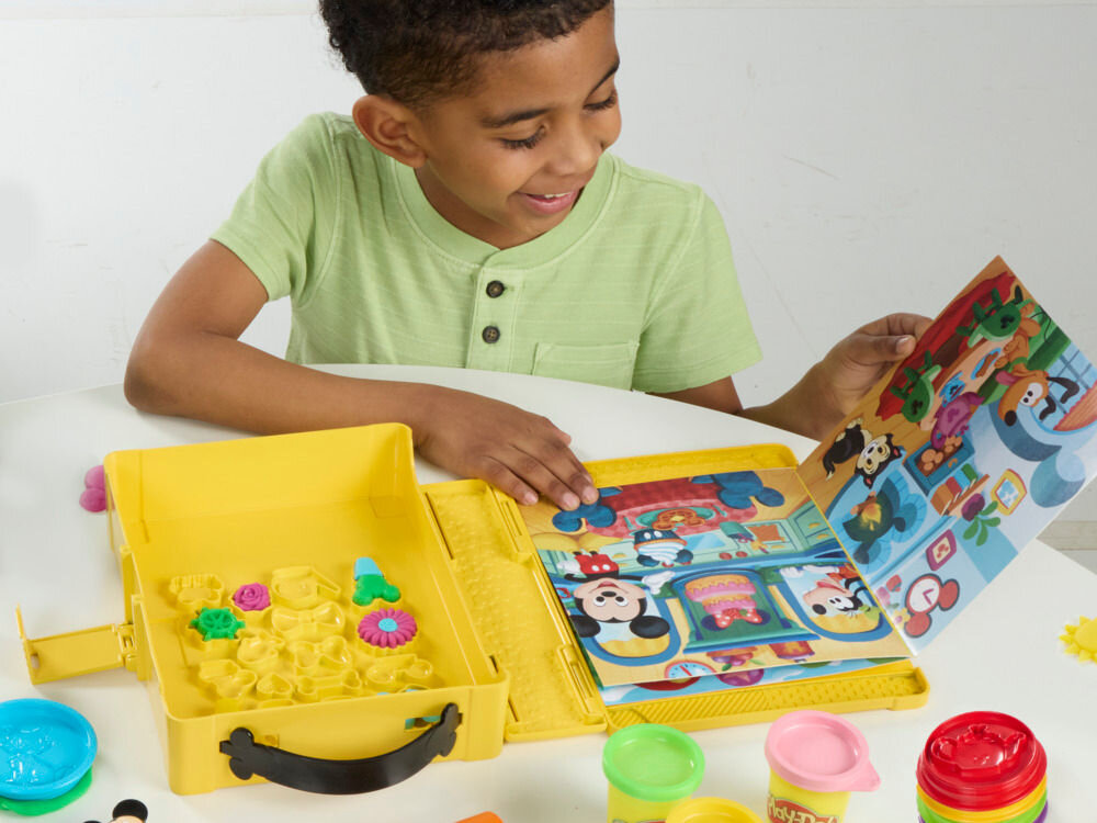 Ciastolina PLAY-DOH Disney Myszka Miki Ciastolinowe Opowieści G23245L0 Chłopiec bawi się zestawem Play-Doh Mickey Mouse Clubhouse, przeglądając kolorową książeczkę z przygodami Disneya obok żółtej walizki pełnej foremek do ciastoliny.