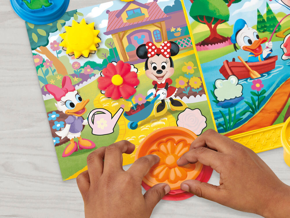 Ciastolina PLAY-DOH Disney Myszka Miki Ciastolinowe Opowieści G23245L0 Zbliżenie na dłonie dziecka tworzące kwiatek z różowej ciastoliny za pomocą pomarańczowej foremki Play-Doh na tle kolorowej planszy z postaciami Daisy i Minnie.