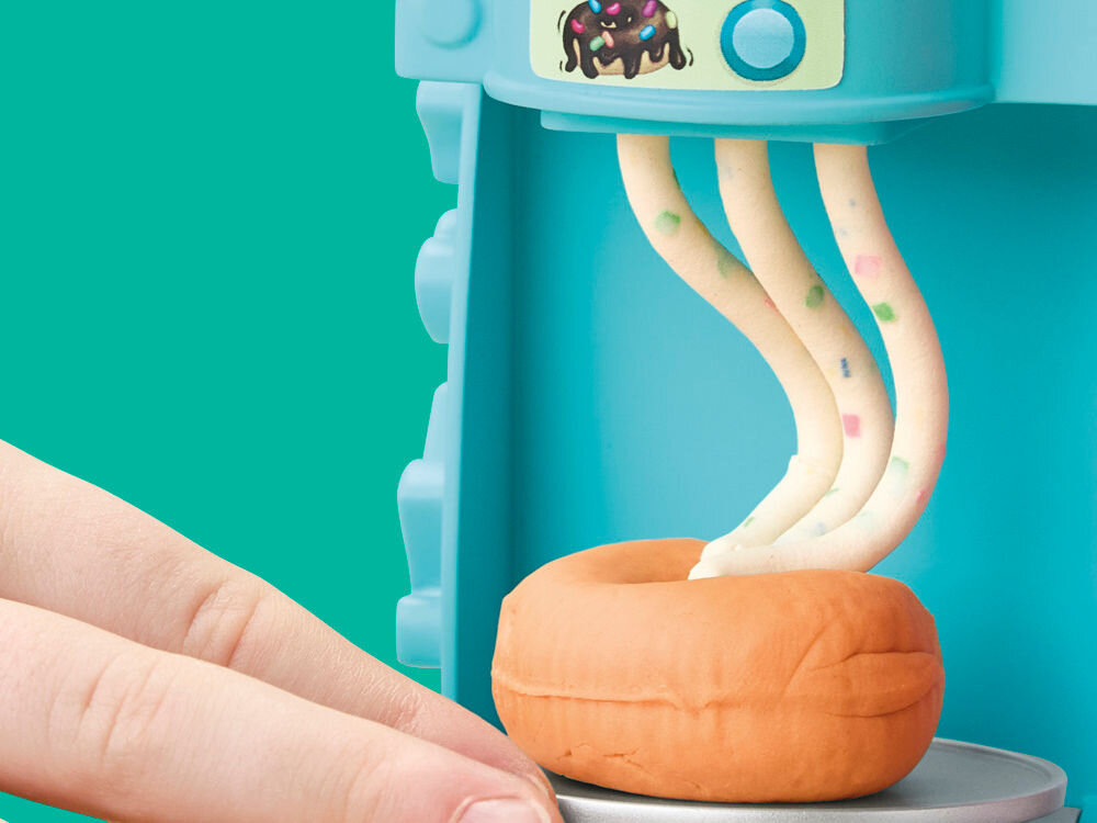 Ciastolina PLAY-DOH Pączkarnia G23085L0 Zbliżenie na dekorowanie pączka Play-Doh: z niebieskiego dozownika wyciskane są cienkie nitki jasnej ciastoliny z kolorowymi drobinkami.