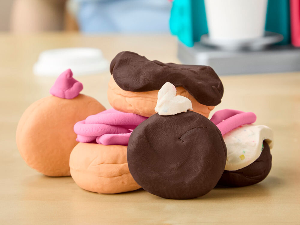 Ciastolina PLAY-DOH Pączkarnia G23085L0 Grupa gotowych pączków i ciastek wykonanych z ciastoliny Play-Doh w kolorach brązowym, pomarańczowym i różowym, ułożona w stos.