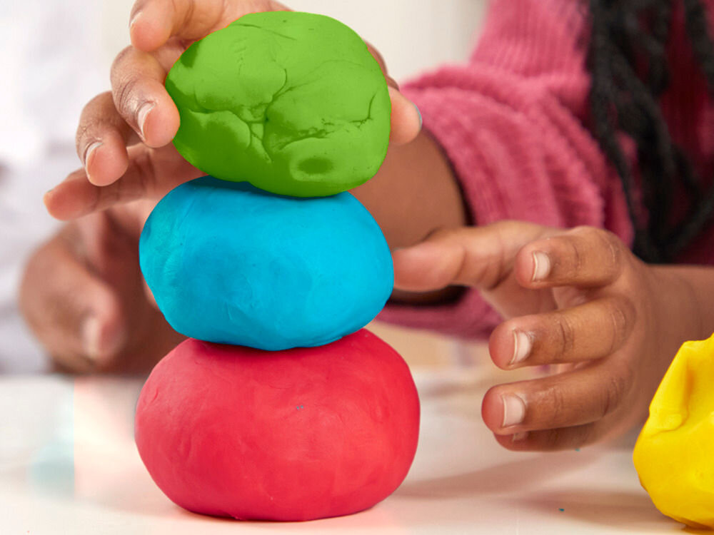 Ciastolina PLAY-DOH Zestaw startowy B11695L0 Zbliżenie na dłonie dziecka układającego pionową wieżę z trzech kulek ciastoliny w kolorach zielonym, niebieskim i czerwonym.