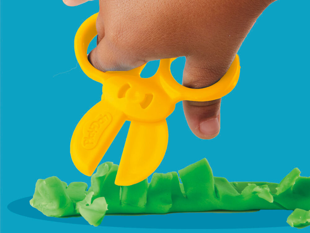 Ciastolina PLAY-DOH Zestaw startowy B11695L0 Dziecko używające żółtych plastikowych nożyczek Play-Doh do wycinania wzorów w podłużnym kawałku zielonej ciastoliny na niebieskim tle.