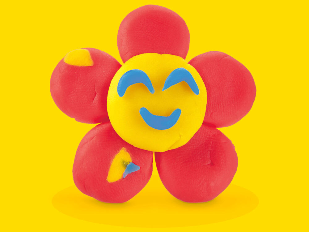 Ciastolina PLAY-DOH Zestaw startowy B11695L0 Uśmiechnięty kwiatek wykonany z ciastoliny z czerwonymi płatkami i żółtym środkiem, na którym naklejono niebieskie oczy i uśmiech, przedstawiony na żółtym tle.