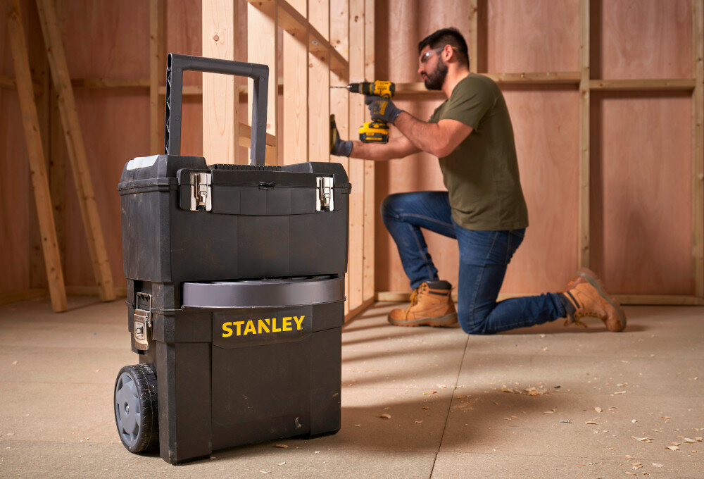 Skrzynka narzędziowa STANLEY 1-93-968 pomieszczenie drewniana podłoga skrzynki ustawione na podłodze w tle klęczy osoba pracuje drewniana ściana drewniana podłoga ruchomy warsztat modułowość mobilność transparent i przechowywanie narzędzi