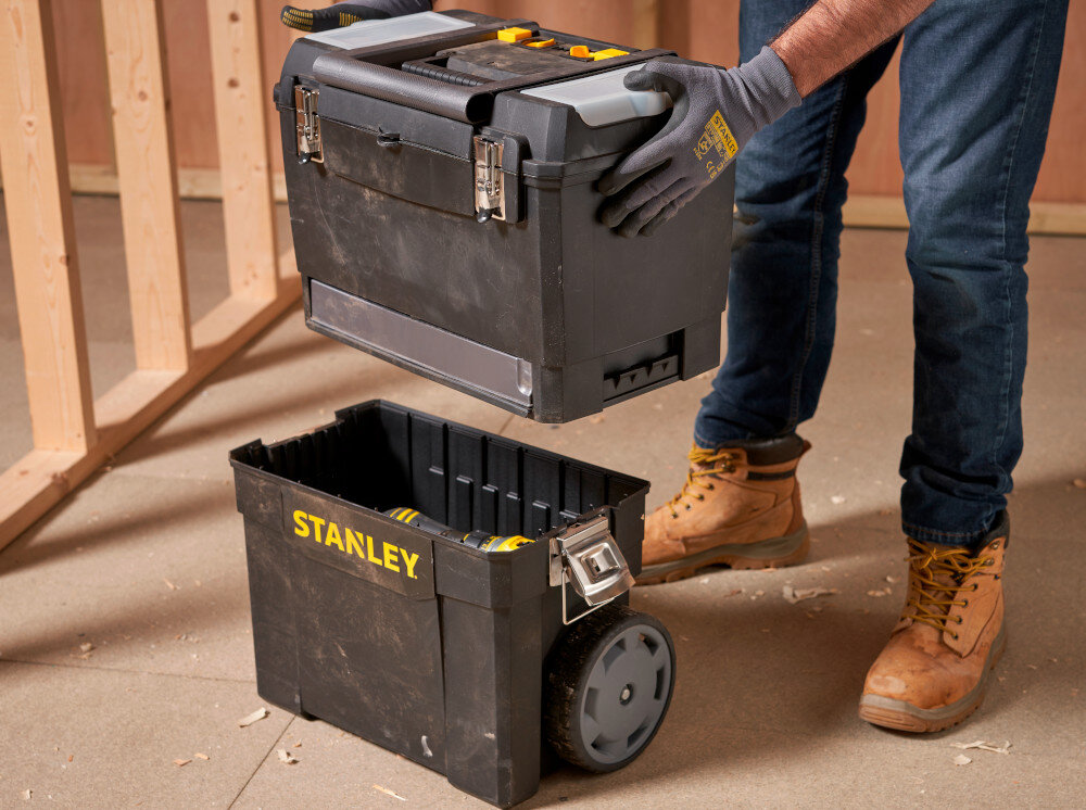 Skrzynka narzędziowa STANLEY 1-93-968 pomieszczenie osoba unosi górną skrzynkę pod skrzynką stoi dolna skrzynka drewniana ściana i podłoga do przechowywania transportu narzędzi konstrukcja modułowa dwie części górna i dolna dopasowany zestaw do rodzaju wykonywanych prac