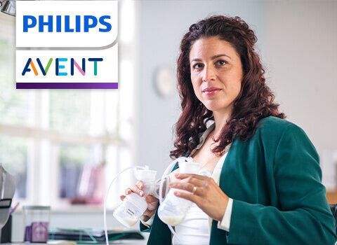 Kobieta trzyma laktator w rękach, w tle widać rozmyte wnętrze pomieszczenia. W lewym górnym rogu znajduje się logo \'PHILIPS AVENT\'.