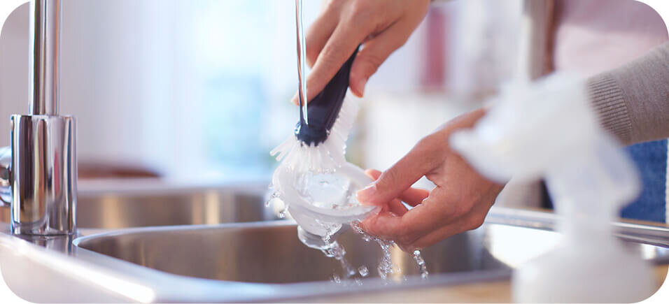 Osoba myje element laktatora pod bieżącą wodą w zlewie, używając szczotki do czyszczenia. W tle widoczny jest rozmyty obrazek innego przedmiotu.