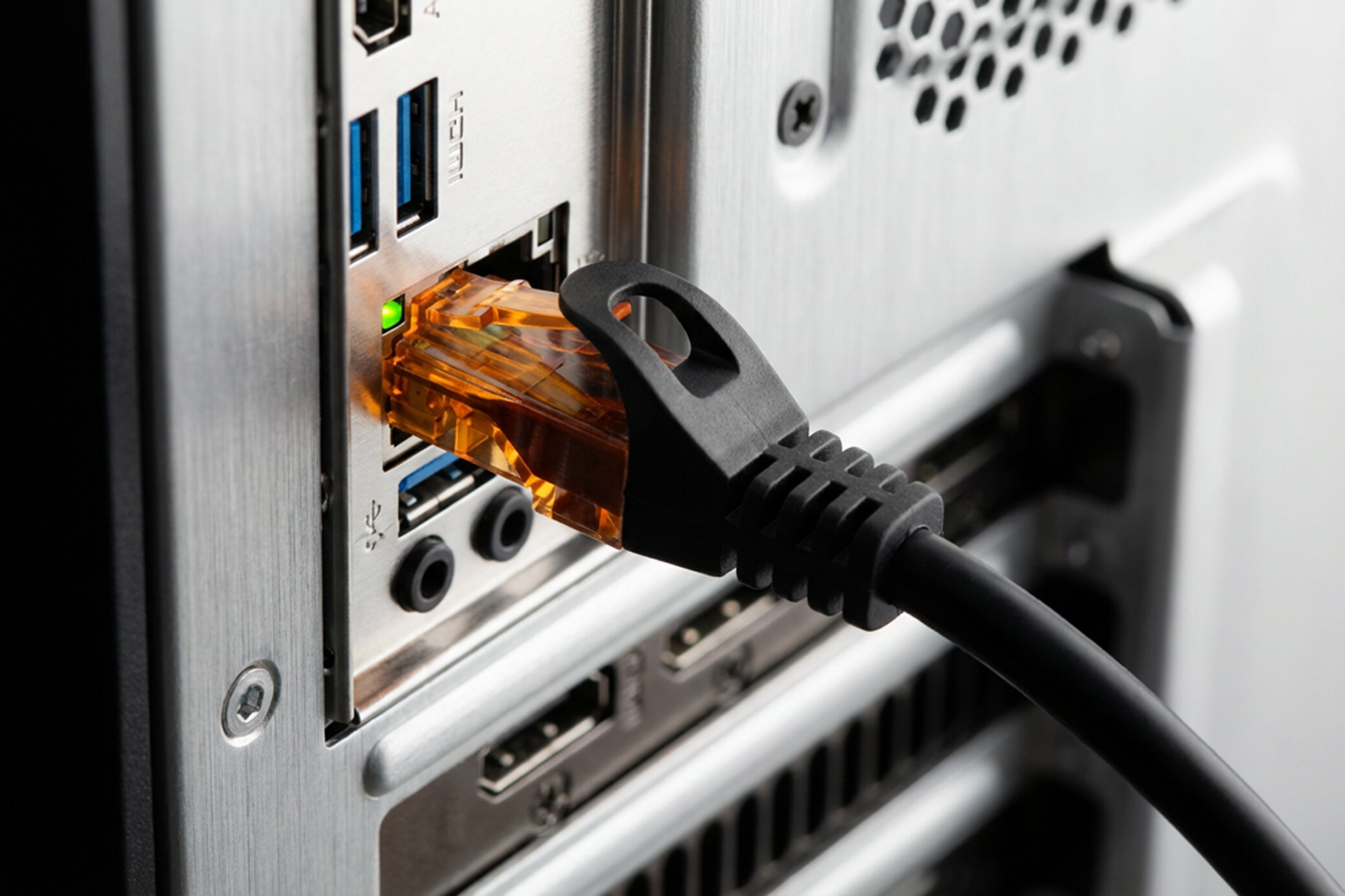 Pomarańczowy kabel RJ-45 podłączony do portu sieciowego na tylnej części komputera. Obok widoczne są porty USB i inne złącza.