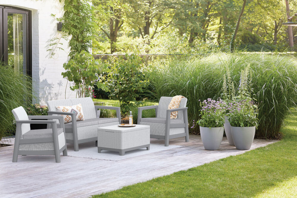 Na drewnianym tarasie znajduje się zestaw mebli ogrodowych w jasnoszarym kolorze, składający się z dwóch foteli, sofy i stolika. W tle widoczna jest bujna roślinność oraz donice z kwiatami.