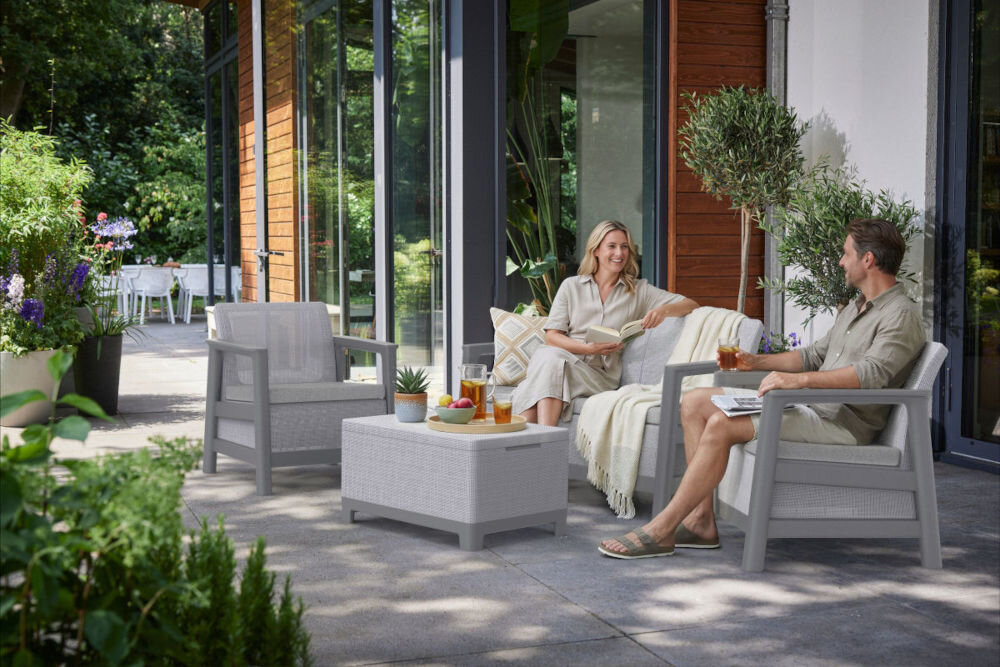 Na tarasie znajdują się jasnoszare meble ogrodowe, w tym dwa fotele, sofa i stolik z napojami oraz owocami. W tle widoczne są rośliny i duże okna budynku.