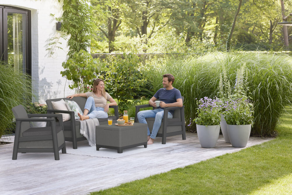 Na tarasie znajdują się grafitowo-szare meble ogrodowe, w tym dwa fotele, sofa i stolik. Obok mebli stoją duże donice z roślinami, a w tle widać zielony ogród.