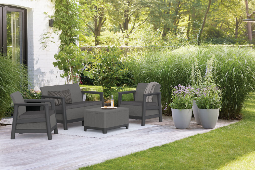 Na obrazie widoczny jest zestaw mebli ogrodowych w grafitowo-szarym kolorze, składający się z dwóch foteli, sofy i stolika. W tle znajdują się zielone rośliny i biała ściana budynku.