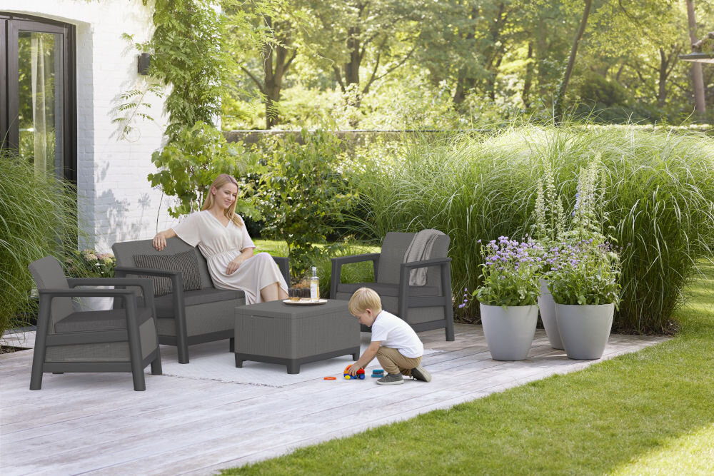 Zestaw mebli ogrodowych w grafitowo-szarym kolorze, składający się z dwóch foteli, sofy i stolika, ustawiony na drewnianym tarasie. W tle widoczne są zielone rośliny i kwiaty w donicach.