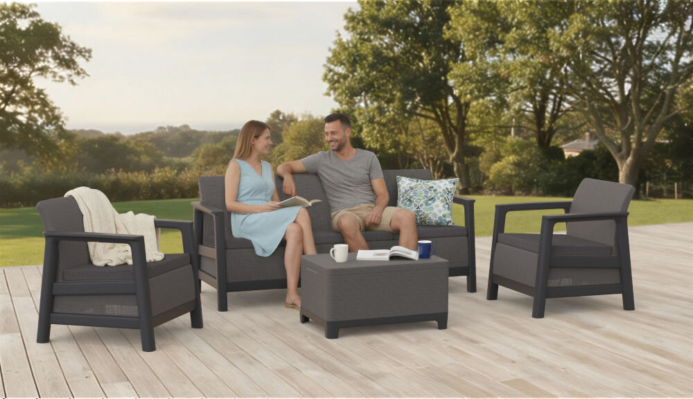 Zestaw mebli ogrodowych w grafitowo-szarym kolorze, składający się z dwóch foteli, sofy i stolika, ustawiony na drewnianym tarasie. W tle widoczny jest zielony trawnik i drzewa.