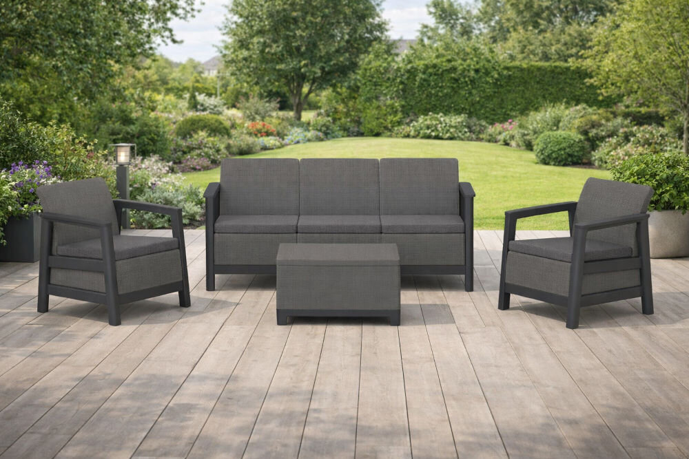 Zestaw mebli ogrodowych składający się z dwóch foteli, sofy i stolika w kolorze grafitowo-szarym, ustawiony na drewnianym tarasie z widokiem na zielony ogród. Meble mają prosty, nowoczesny design z poduszkami na siedziskach i oparciach.