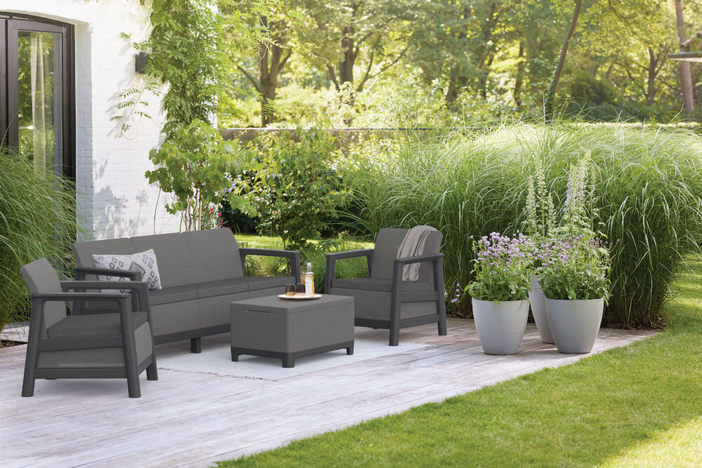 Na drewnianym tarasie znajduje się zestaw mebli ogrodowych w kolorze grafitowo-szarym, składający się z sofy, dwóch foteli i stolika. W tle widoczne są wysokie trawy i rośliny w donicach.