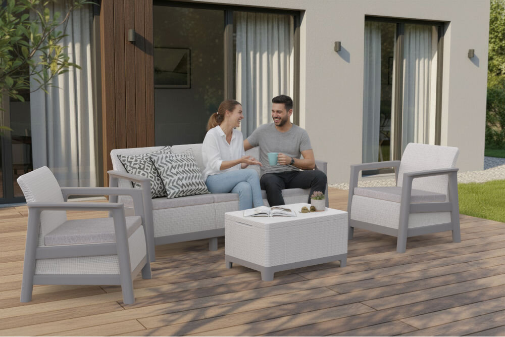 Na drewnianym tarasie przed domem znajduje się zestaw mebli ogrodowych w jasnoszarym kolorze, składający się z dwóch foteli, sofy i stolika. Na sofie siedzą dwie osoby, rozmawiając i trzymając kubki.