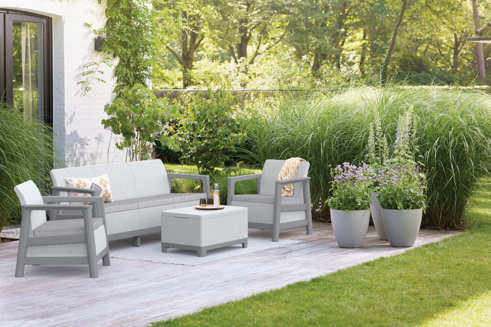 Na zdjęciu znajduje się zestaw mebli ogrodowych w jasnoszarym kolorze, składający się z sofy, dwóch foteli i stolika. Meble ustawione są na drewnianym tarasie, otoczone zielenią i donicami z roślinami.