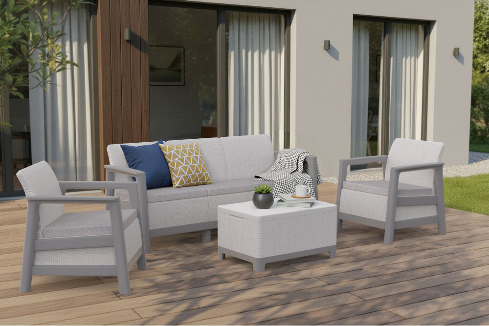 Zestaw mebli ogrodowych w jasnoszarym kolorze, składający się z dwóch foteli, sofy i stolika, ustawiony na drewnianym tarasie przed domem z dużymi oknami. Na sofie znajdują się poduszki w różnych kolorach, a na stoliku stoi doniczka z rośliną i filiżanka.