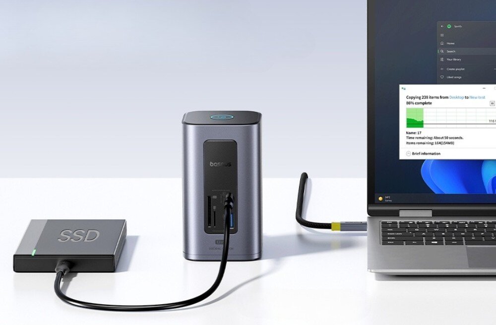 Stacja dokująca BASEUS NU1 Air Spacemate Zbliżenie na stację dokującą podłączoną kablem do laptopa, po lewej stronie zewnętrzny dysk SSD z napisem 'SSD', na ekranie laptopa widoczny pasek postępu kopiowania plików