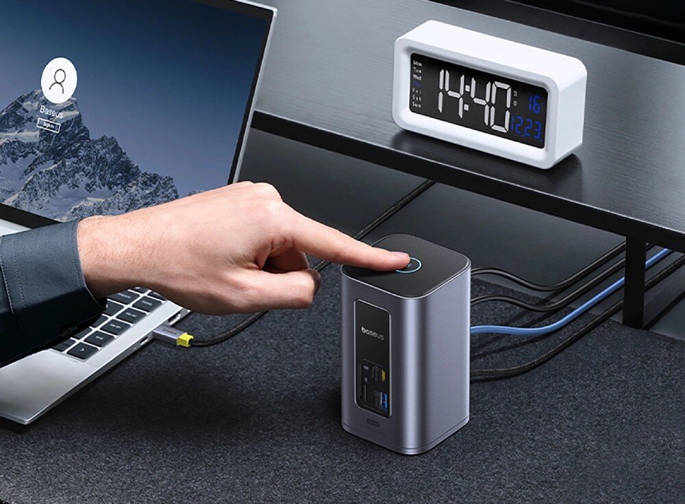 Stacja dokująca BASEUS NU1 Air Spacemate Pionowa stacja dokująca w szarej obudowie stojąca na ciemnym biurku obok laptopa, w tle biały zegar cyfrowy i monitor
