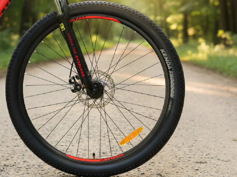 Rower górski MTB TATRA Overland U21 29 cali Czerwony las droga zbliżenie na przednie koło opony marki KENDA w rozmiarze 29 cali głęboki bieżnik skutecznie wspiera jazdę ułatwia utrzymanie stabilnego toru jazdy aluminiowe obręcze stalowe piasty marki Ciclo Sport regularne sprawdzanie ciśnienia powietrza wartość na bocznej części opony