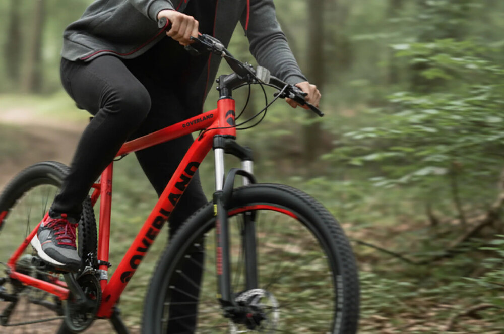 Rower górski MTB TATRA Overland U21 29 cali Czerwony las osoba jedzie na rowerze po drodze leśnej zbliżenie na przednie koło i amortyzator konstrukcja hardtail z przednim amortyzatorem Ciclo Sport o skoku 100 mm stalowa, sprężynowa konstrukcja z aluminiową koroną funkcja LOCKOUT można zablokować pracę amortyzatora na gładkiej nawierzchni