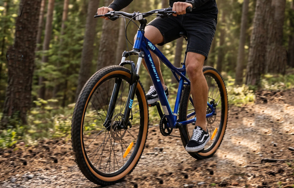 Rower górski MTB SUN BABY Verdant Shred D03.03.09.4.1 U17 27.5 cala Niebieski las ścieżka osoba jedzie na rowerze po leśnej ścieżce rower typu hardtail przedni amortyzowany widelec SBB ogranicza przenoszenie nierówności dłonie i ramiona są mniej narażone na odczuwanie wstrząsów