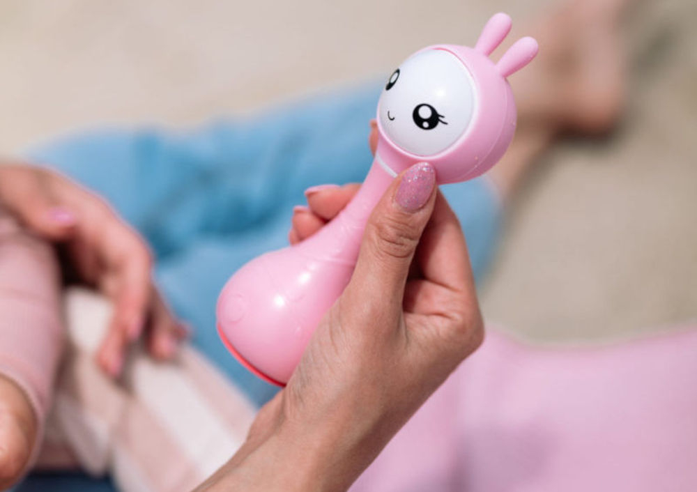 Zabawka edukacyjna ALILO Smarty Bunny R1 Niebieski delikatne światło nocne, które pomaga w budowaniu poczucia bezpieczeństwa podczas 
					zasypiania i sprzyja spokojnemu wypoczynkowi, miękkie, świecące uszy króliczka, subtelne, zmieniające się światło, 7 kolorów,
					produkt w wersji różowej w kobiecej dłoni