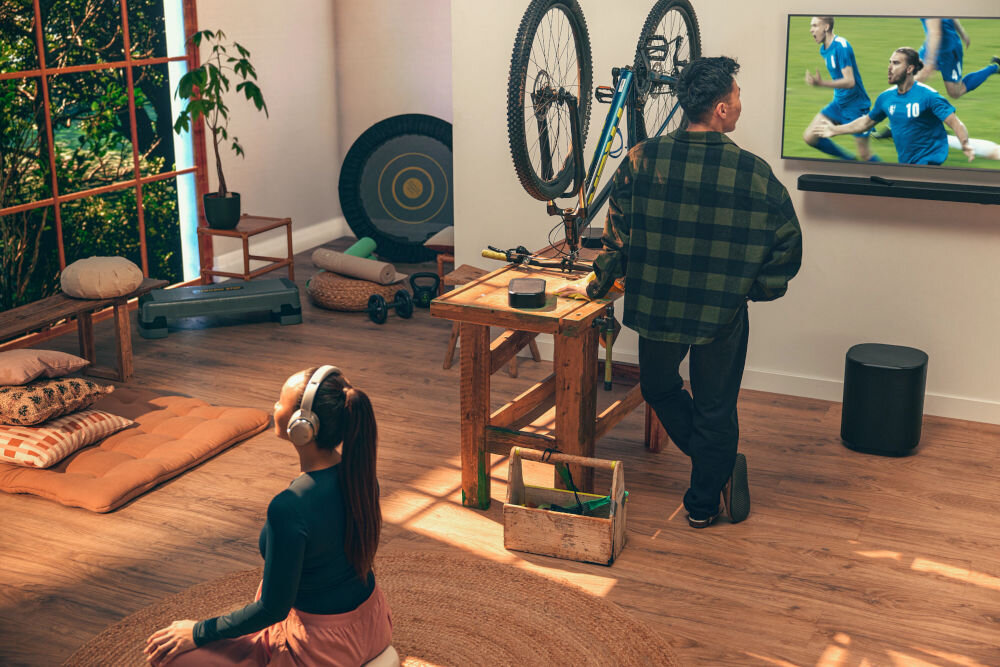 Soundbar JBL BAR 1000 MkII 960 W moc całkowita wydajność kino domowe, osoba stojąca przed telewizorem oglądająca mecz, osoba siedzaca po turecku w słuchawkach na dywanie