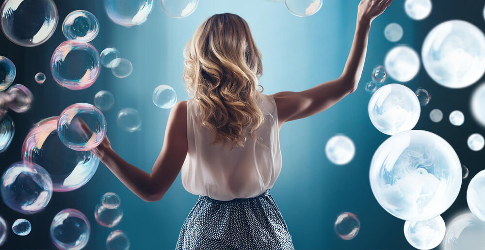 Osoba stoi tyłem w otoczeniu dużych, przezroczystych baniek mydlanych na tle niebieskiego światła. Bańki mają różne rozmiary i unoszą się w powietrzu.