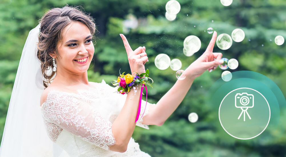 Osoba w białej sukni ślubnej stoi na tle zieleni, unosząc ręce w stronę unoszących się baniek. W prawym dolnym rogu znajduje się ikona aparatu fotograficznego na statywie.