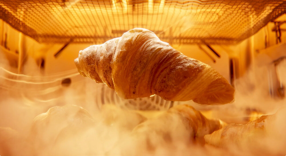 Piekarnik DREAME Combi OZ60Pro Zbliżenie wnętrza piekarnika z unoszącymi się croissantami i widoczną parą, podkreślającą pieczenie w wysokiej temperaturze. Ciepłe, złote światło eksponuje strukturę wypieków równomierne pieczenie 10 trybów para Steam Assist 3D Hot Air soczyste wnętrze złocista skórka puszyste wypieki