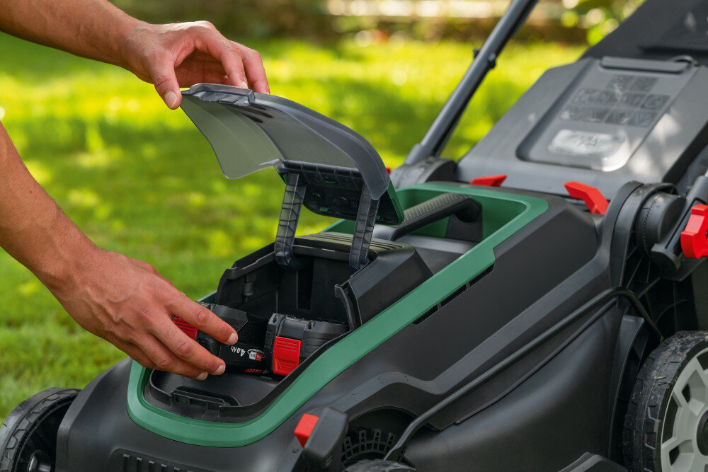 Osoba wkłada akumulator do komory kosiarki akumulatorowej, której pokrywa jest otwarta. Kosiarka znajduje się na trawie, a w tle widoczne są rozmyte elementy zieleni.