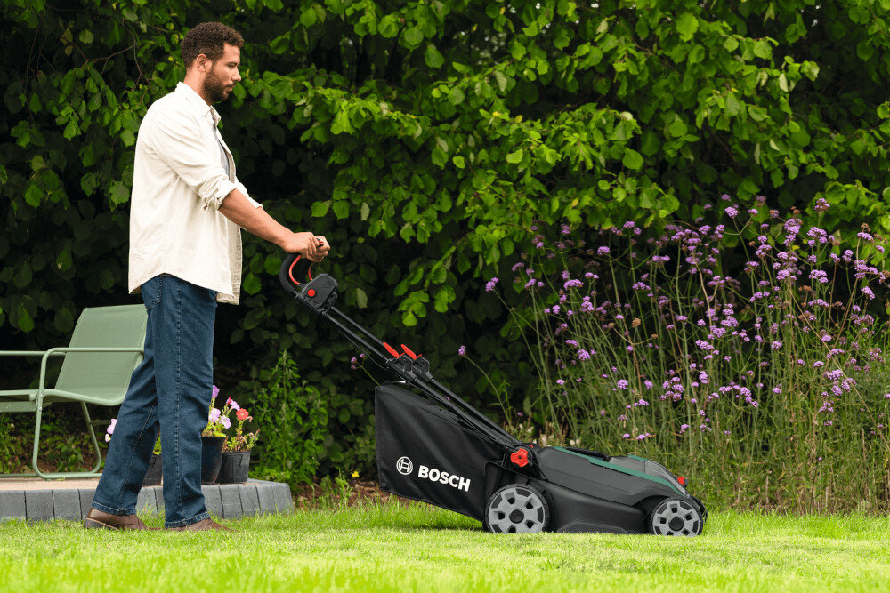 Osoba pcha kosiarkę akumulatorową z widocznym logo BOSCH na trawniku, w tle zielone krzewy i fioletowe kwiaty.