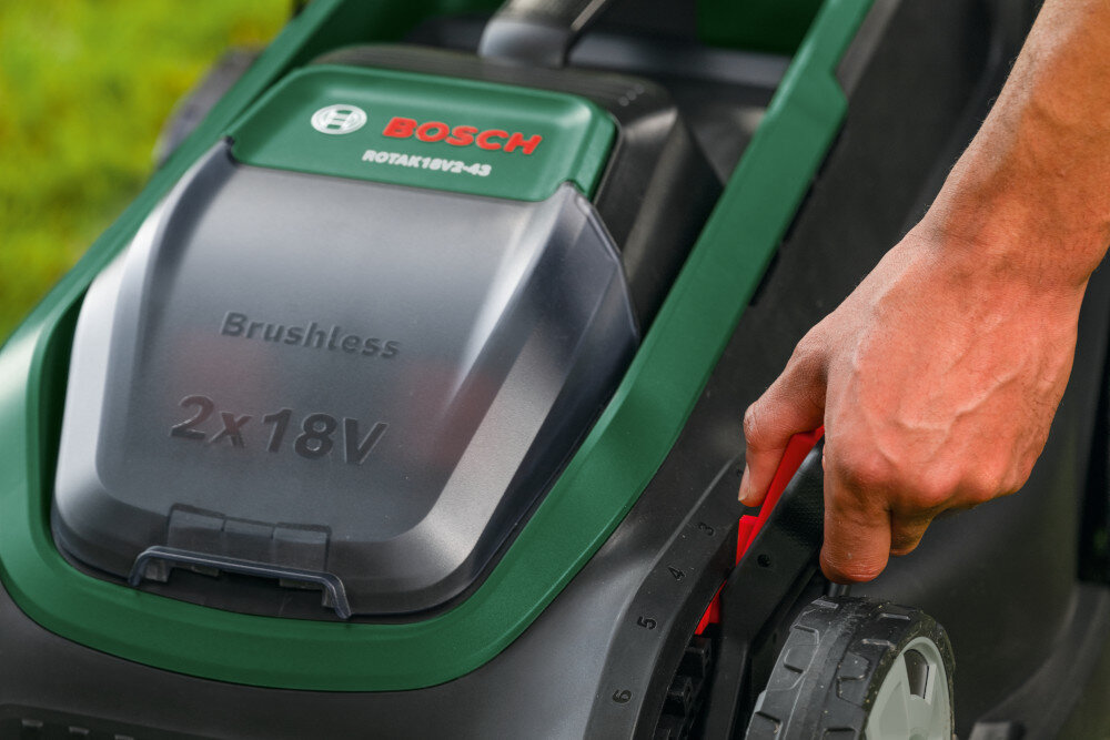 Na zdjęciu widoczna jest część kosiarki z logo BOSCH oraz napisami \'ROTAK 18V2-43\', \'Brushless\', \'2x18V\'. Osoba reguluje wysokość kosiarki za pomocą czerwonego uchwytu przy kole.