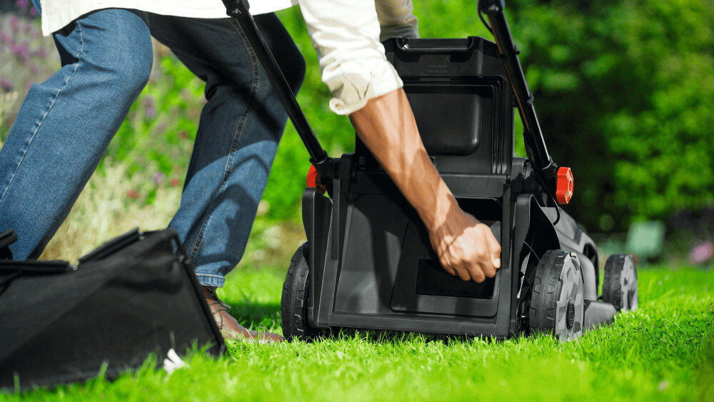 Osoba wkłada pojemnik do tylnej części czarnej kosiarki stojącej na trawie. Kosiarka ma duże koła i czerwone elementy przy uchwytach.