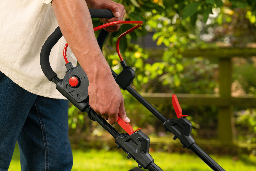 Osoba trzyma uchwyt kosiarki z czerwonymi elementami sterującymi. W tle widoczne są zielone liście i drewniany płot.