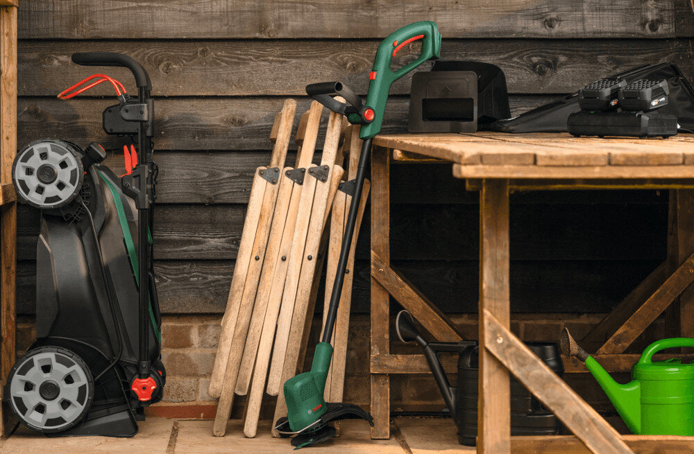 Na obrazie widać kosiarkę akumulatorową złożoną pionowo obok drewnianego stołu, na którym znajdują się narzędzia ogrodowe i akcesoria. W tle widoczna jest drewniana ściana oraz kilka drewnianych uchwytów.