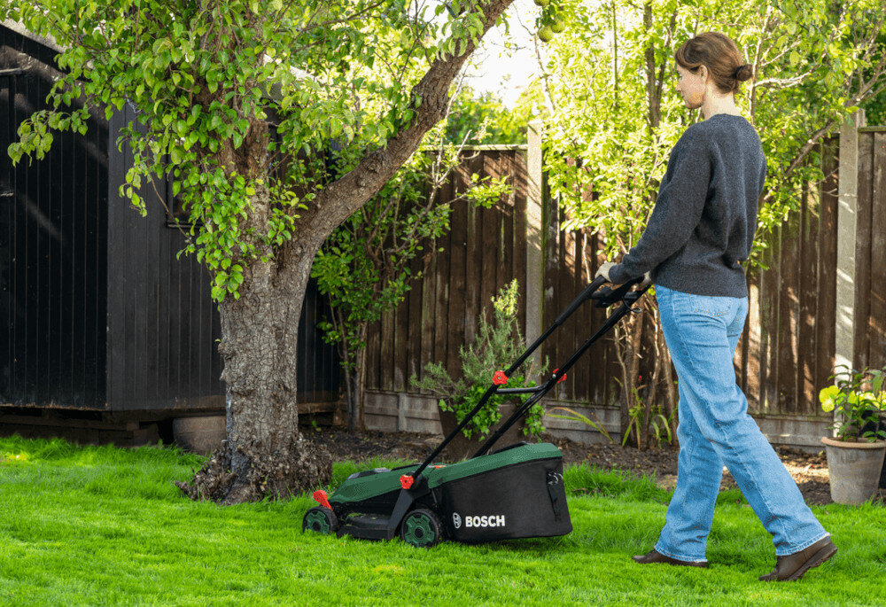 Osoba kosi trawnik za pomocą zielonej kosiarki z widocznym logo BOSCH. W tle znajduje się drzewo i drewniany płot.