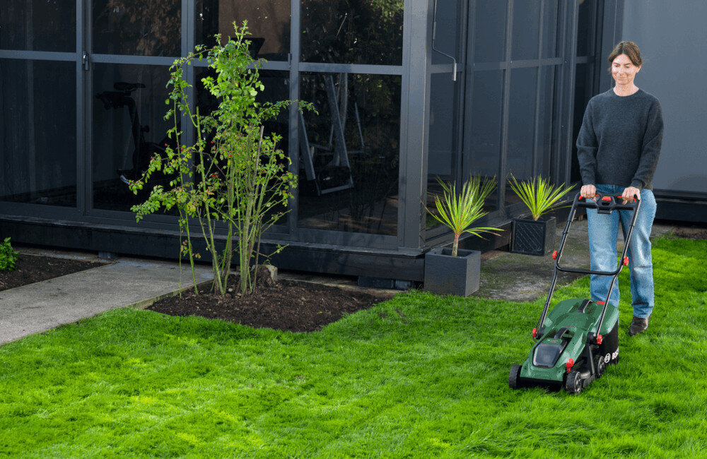 Osoba kosi trawnik za pomocą zielonej kosiarki akumulatorowej przed nowoczesnym budynkiem z dużymi oknami. W tle widoczne są rośliny w donicach oraz krzewy.