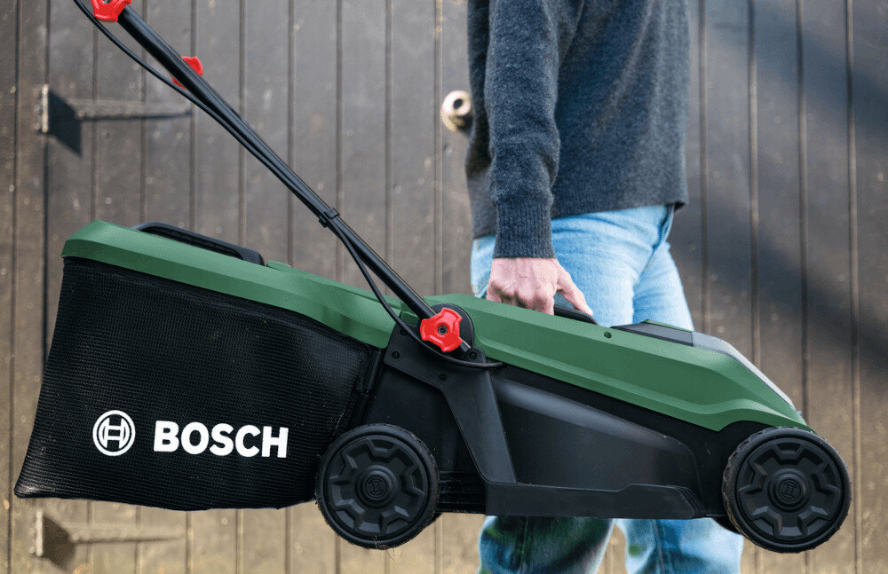 Osoba trzyma kosiarkę akumulatorową w kolorze zielonym i czarnym z widocznym logo BOSCH na koszu. W tle znajduje się drewniana ściana.