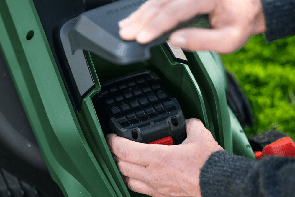 Osoba wkłada akumulator do zielonego urządzenia ogrodowego. Widoczna jest część mechanizmu z czarnym i czerwonym elementem.