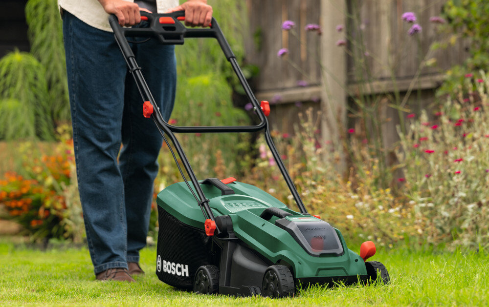 Osoba pcha zieloną kosiarkę akumulatorową z widocznym logo BOSCH po trawniku. W tle widać kwiaty i drewniane ogrodzenie.