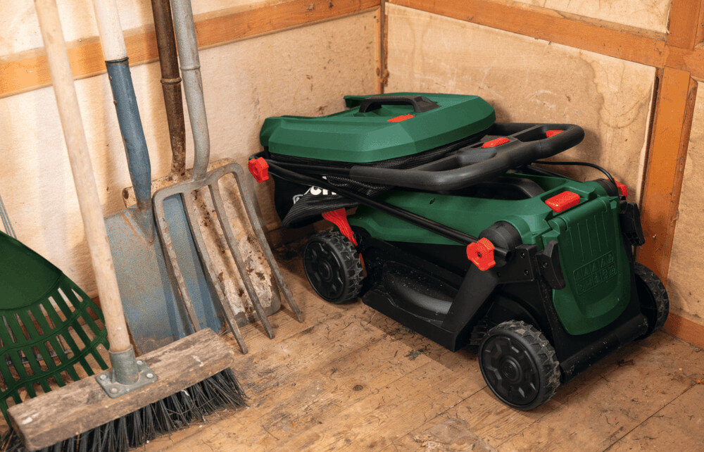 W rogu pomieszczenia stoi złożona kosiarka akumulatorowa w kolorze zielonym z czarnymi i czerwonymi elementami. Obok niej znajdują się narzędzia ogrodowe, takie jak grabie i łopaty.