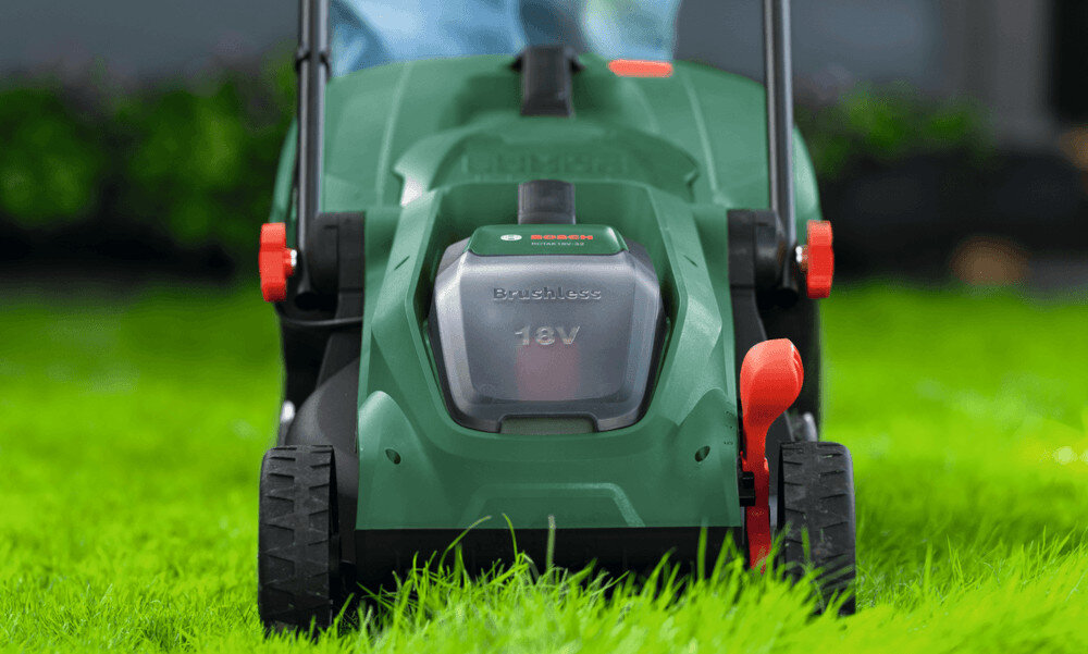 Zielona kosiarka z widocznym napisem \'Brushless 18V\' na obudowie, stojąca na trawie. Na kosiarkę nałożone są czerwone elementy sterujące.
