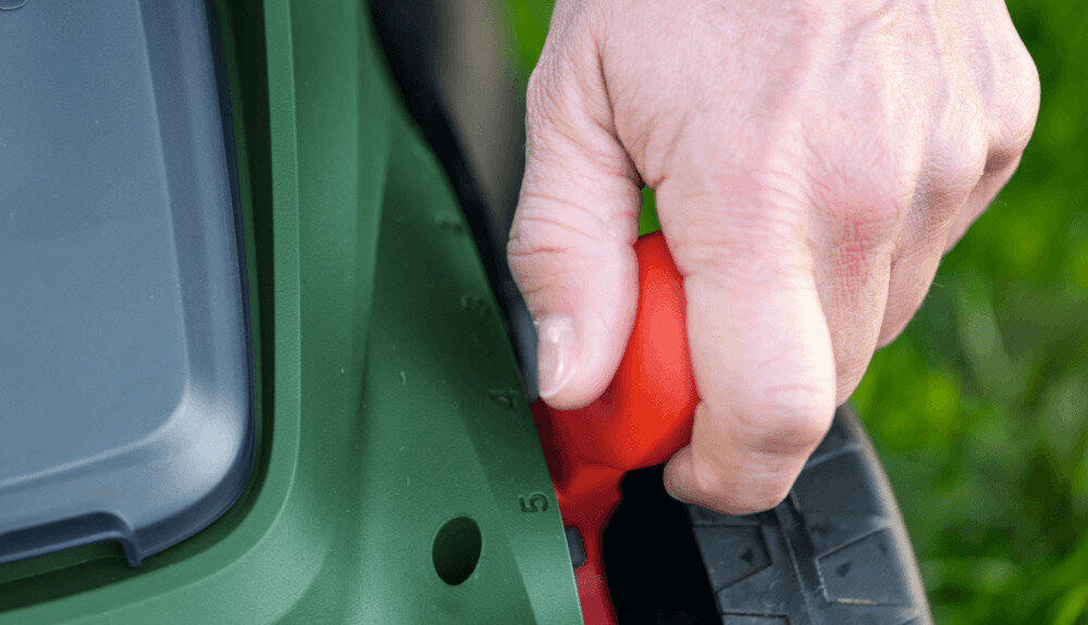 Zbliżenie na rękę regulującą wysokość kosiarki za pomocą czerwonego pokrętła na zielonej obudowie. Obok widoczne są oznaczenia wysokości w postaci cyfr.