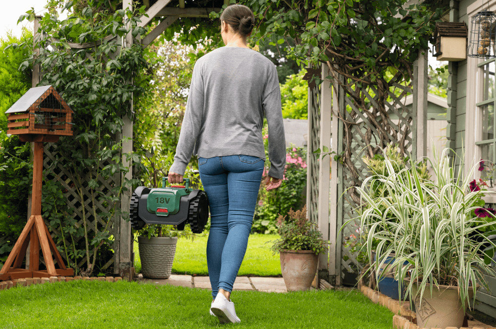 Robot koszący  BOSCH Visimow 18V-100 06008E1001 Bez przewodu ograniczającego ogród osoba widoczna tyłem niesie w dłoni robot krzewy kwiaty rabaty dom silnik bezszczotkowy dłuższa żywotność wyższa wydajność urządzenie może pracować dłużej bez spadków mocy bardziej efektywne koszenie mniejsza potrzeba serwisowania niski poziom hałasu wynoszący 52 dB cichsza praca