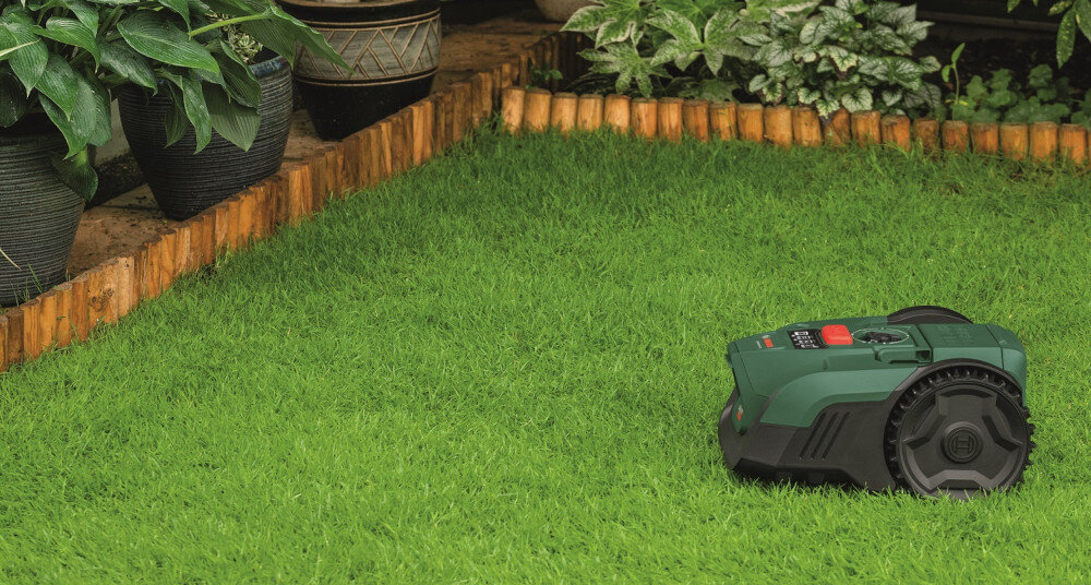 Robot koszący  BOSCH Visimow 18V-100 06008E1001 Bez przewodu ograniczającego ogród trawnik robot stoi na trawniku widok lekko z góry rabaty kwiatowe technologia SmartVision wykrywanie powierzchni trawiastych i nietrawiastych bardziej precyzyjne koszenie sam rozpoznaje, gdzie powinien pracować trawnik jest pielęgnowany równomiernie otoczenie pozostaje nienaruszone