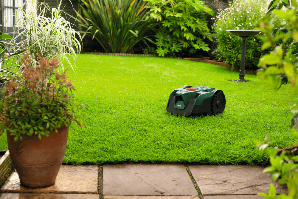 Robot koszący  BOSCH Visimow 18V-100 06008E1001 Bez przewodu ograniczającego ogród trawnik robot stoi na trawniku w tle krzewy fontanna kwiaty system trzech obrotowych noży szerokość cięcia 16 cm równomierne skracanie trawy na całej powierzchni dokładnie przycina źdźbła efekt jest bardziej jednolity praca przebiega bez wysiłku